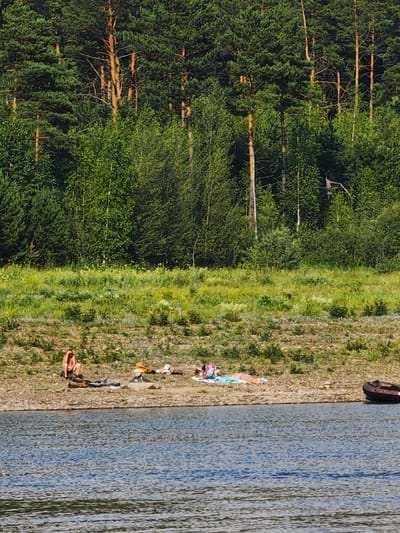 The image depicts a waterfront scene on a sunny day, featuring a densely wooded shoreline of pine and deciduous trees. In the foreground, the blue water of a river or lake is visible, with ripples indicating light movement. Along the pebble and dirt embankment, several individuals are sunbathing. On the left, a person is seated, appearing to be resting. Further to the right, other individuals are lying on towels or directly on the ground. Clothing items, including what appears to be an orange life vest, are scattered around the sunbathers. In the far right of the image, a dark-colored inflatable boat is partially submerged at the water's edge. The environment suggests a natural recreational area, potentially near the city of Prutovaya, Russia, given the typical landscape of the region.