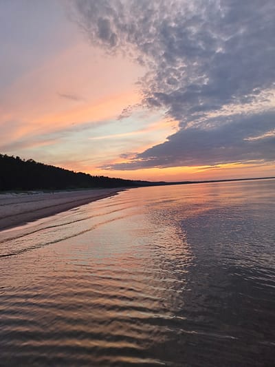This image depicts a serene beach scene in Apšuciems, Latvia, during sunset. The sky is a canvas of soft pastel colors, with hues of pink, orange, and lavender blending into a soft grey towards the top, where scattered clouds are visible. A dark line of trees forms a dense forest along the coastline, bordering a sandy beach. The gentle waves of the Baltic Sea lap at the shore, reflecting the warm colors of the sky. The water is calm, with subtle ripples creating a shimmering effect on the surface. A single person is faintly visible walking on the beach, adding a sense of scale to the vast landscape. The overall impression is one of peaceful tranquility and natural beauty.