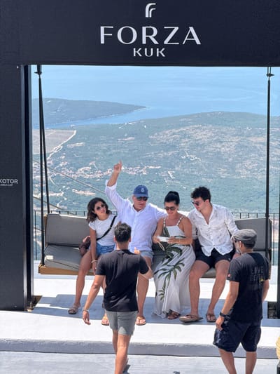 This media file shows a group of six people standing and sitting on a white, outdoor platform. The platform appears to be elevated, offering a panoramic view of a bay with a town nestled along its shores, surrounded by lush green hills. The water is a clear blue, and the sky is bright and sunny, suggesting a warm, fair-weather day. The time of day appears to be afternoon, indicated by the sunlight and shadows.
The individuals are casually dressed in summer attire, including t-shirts, shorts, and dresses. Some are looking at the view, while others are interacting with each other. One person is pointing towards the landscape. The atmosphere seems relaxed and convivial, with the group enjoying the scenic vista.
A prominent black sign with white lettering is visible on the left side of the frame, displaying the text "FORZA KUK". This, combined with the mountainous coastal landscape, strongly suggests the location is Kuk, Montenegro. Another partially visible sign at the bottom reads "KOTOR TABLE CAP".
The people are positioned in various ways: some standing, some sitting on lounge chairs. The scene is dynamic, capturing a moment of shared experience and appreciation for the beautiful surroundings. The overall impression is one of leisure and sightseeing in a picturesque coastal town.