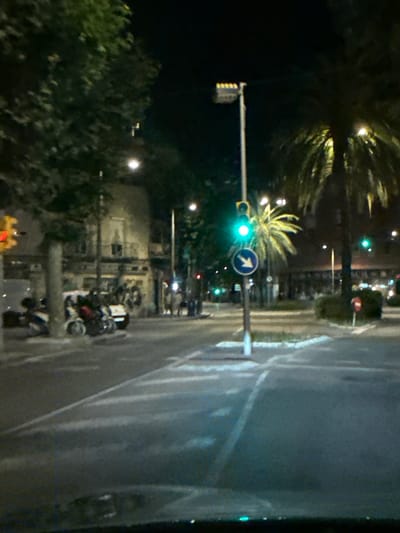 The image depicts a night-time street scene in l'Hospitalet de Llobregat, Spain, possibly near the intersection of Rambla Just Oliveras and Carrer de Llobregat. A traffic light is illuminated with a green signal, indicating that traffic can proceed. A blue circular sign with a white arrow pointing to the right is visible beneath the traffic light, suggesting a mandatory right turn. Several parked motorcycles and scooters are lined up on the left side of the street, some with their lights on. A palm tree with illuminated fronds is prominent on the right side of the image, alongside other vegetation. Buildings with darkened windows are visible in the background, some appearing to be residential or commercial establishments. Streetlights and other ambient lights illuminate the road and sidewalks, creating reflections on the wet pavement, which indicates recent rain or a damp surface. The overall atmosphere is that of a quiet urban street at night.
