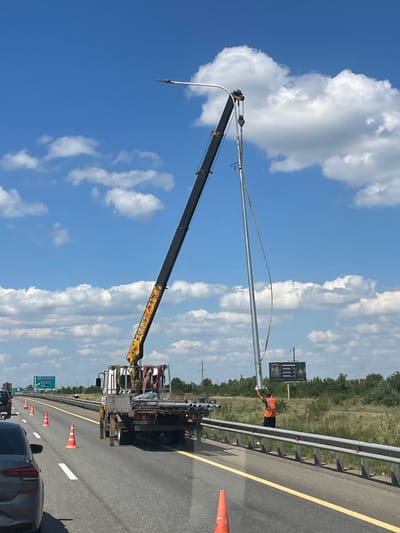On a highway in Molkino, Russia, a crane truck is installing a new street light pole. The truck is positioned on the side of the road, with its crane extended to lift the pole. A worker in an orange vest is guiding the pole into place, while another person is visible inside the truck's cab. Orange traffic cones are placed around the work area to alert other drivers. In the background, there is a billboard and a tree line. The sky is blue with scattered clouds.