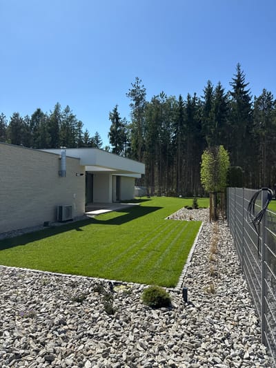The image depicts a modern house and its landscaped yard in Wiązowna, Poland, under a clear blue sky. The house is rectangular, with a light-colored brick exterior, a flat white roof, and large windows. An air conditioning unit is visible on the side of the house. A trimmed green lawn stretches from the house to a gray gravel bed, which is bordered by a metal fence. Trees are visible in the background.