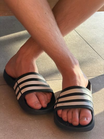 A close-up shot shows a person's legs and feet, positioned as if sitting with legs crossed. The feet are wearing black Adidas slides with white stripes. The individual has light skin and visible leg hair. The background is a tiled floor, with sunlight casting shadows. The scene is set in Khotianivka, Ukraine.