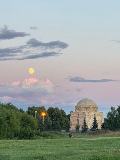 In the city of Bolgar, Russia, a circular stone mausoleum with a dome stands in the foreground against a twilight sky with scattered pink and gray clouds. A full, luminous moon hangs in the sky, situated above soft pink clouds. The mausoleum is surrounded by a grove of dark green coniferous trees and is enclosed by a black metal fence. To the left of the mausoleum, a tall lamppost with two orange lights illuminates the area. In the middle ground, a person is walking in a grassy field, facing away from the camera and towards the mausoleum. The field is covered in green grass and various plants. The background consists of dense trees and a faint, pale blue sky.