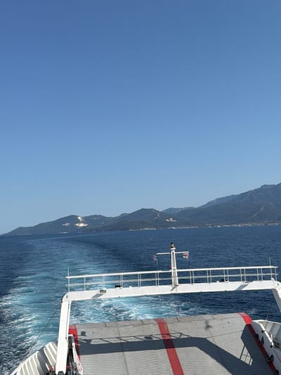 This is a vertical, medium-shot photograph taken from the deck of a ferry, looking out towards a coastline with mountains. The sky is a clear, bright blue, indicating a sunny day. The sea is a deep blue, with white wakes trailing behind the ferry.

The ferry's structure is visible on the right side of the image, with grey metal siding and red stripes. A white metal railing runs along the side of the ferry, extending into the water. Two flags are visible on a pole attached to the ferry: one is a white flag with a red cross, and the other appears to be the Greek flag. The shoreline, characterized by lush green mountains and some scattered buildings, is visible in the distance on the left.

The overall scene suggests a leisurely journey on a ferry under clear skies. The lighting indicates it is daytime. The location is Keramoti, Greece, which aligns with the mountainous coastline and the presence of the Greek flag. There is no visible text in the image.