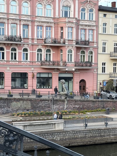 The image shows a section of the Brda River canal in Bydgoszcz, Poland, with a view of buildings and the adjacent promenade. The buildings are multi-story, with pink and cream facades featuring arched windows, some with decorative balconies. A sculpture of a nude man crossing the river on a tightrope is a prominent element of the canal-side view. Pedestrians walk along the paved promenade beside the canal, which is lined with flowerbeds and a brick wall. A metal railing is visible in the foreground, likely part of a bridge or viewing platform. A "P" parking sign is seen in the background.