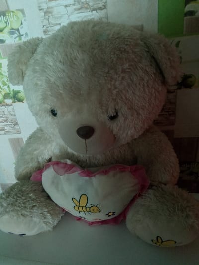 A light-colored teddy bear sits on a white surface, holding a heart-shaped pillow with a bee design. The bear is facing the camera, with its head slightly tilted. The background is a wall with a brick pattern and some green accents, possibly wallpaper. There are no people visible in the image. The lighting is dim, with a strong highlight on the bear's fur. The location is stated as Изыхские Копи, Russia, but the image itself does not contain any visual cues to confirm this location.