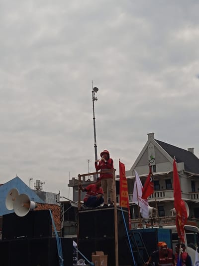 The image was taken in Yogyakarta, Indonesia, at the coordinates -7.783, 110.367. Two individuals are standing on a stage made of wooden planks and large speaker boxes. One is speaking into a microphone. Both individuals are wearing red shirts and reflective vests. Behind them are red and white flags. The stage is in front of a building with a light gray facade, a green roof, and white window frames. On the left side of the image, there is a large speaker system with two blue speaker horns. In the background, a tall metal pole with a satellite dish and other equipment at the top can be seen. There are also various wires and cables visible in the image. The sky is overcast and gray.