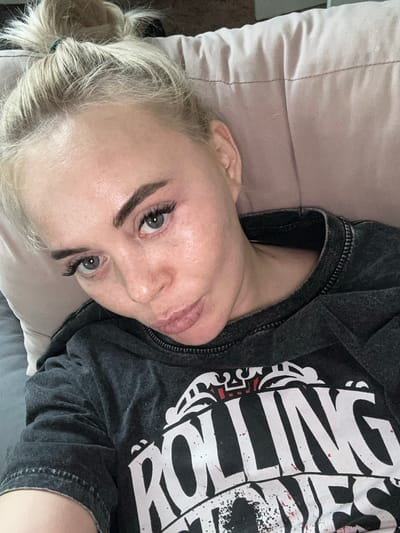 A woman with blonde hair pulled back in a ponytail lies on a bed with a light pink pillow. She is wearing a dark gray t-shirt with a white design. The woman is looking up at the camera. No specific details indicate this is in the city of Высокий, Russia.