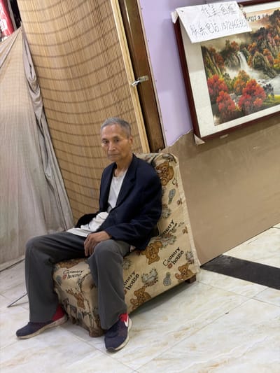 In Guta, China, an elderly East Asian man sits on a patterned armchair. He is wearing a dark blue blazer over a white t-shirt and grey trousers. His feet are clad in blue and red athletic shoes. The armchair's fabric is cream-colored and features a repeating pattern of teddy bears and the words "Country House". Behind the man, a woven bamboo screen forms the backdrop, partially obscured by a light-colored drape on the left. To the right, a framed landscape painting depicting a waterfall amidst autumn foliage hangs on a wall. A white piece of paper with handwritten Chinese text and a phone number is affixed to the painting's frame. The floor is tiled with a light-colored, marbled pattern.