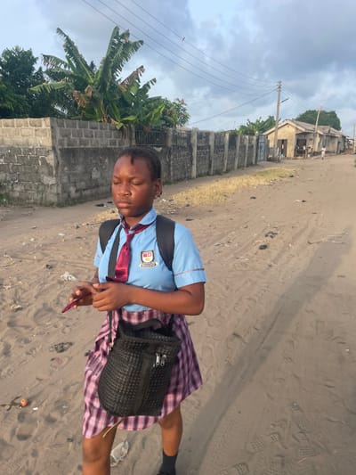 A young female walks on a sandy dirt road in Mosafejaw, Nigeria, under a partly cloudy sky. The subject, appearing to be a student, is centrally framed, facing slightly right, with her eyes closed. She has dark skin and braided hair. She wears a light blue short-sleeved school uniform shirt with red trim and a visible crest on the left chest, a dark red tie, and a red and white plaid skirt. A black backpack is worn on her shoulders, and a small black woven shoulder bag hangs from her left shoulder. She holds a dark red rectangular object, likely a smartphone, with her hands clasped at waist level. To the left, a grey concrete block wall is partially obscured by green vegetation, including banana trees. In the background on the right, a series of single-story, light-colored buildings with corrugated roofs line the road, alongside utility poles and overhead lines. A distant, indistinct human figure is present near these structures. The road surface exhibits tire tracks, footprints, and scattered debris. Long shadows extend towards the right side of the frame.