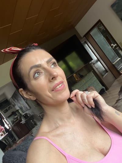 A woman with dark hair and fair skin is seen from a low, angled perspective. She wears a pink top and a striped headband. She is holding a lock of her dark hair in her hand, which features a silver ring and a heart tattoo on the wrist. Her expression is pleasant, and she appears to be looking upwards. The background shows a room with a television, furniture with glass displays, and wooden walls. The location cannot be determined as being within the city of Konz, Germany based on this image alone.