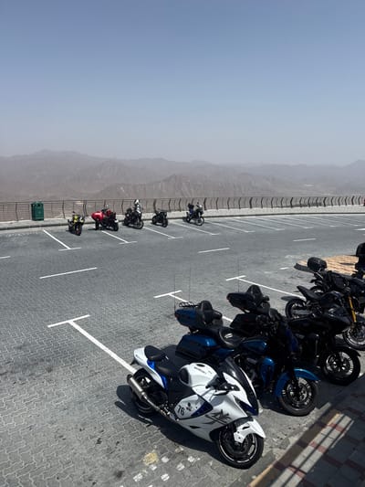 The image shows a parking lot in Khor Fakkan, United Arab Emirates, filled with a variety of motorcycles. The motorcycles are parked in designated spots marked by white lines. A fence runs along the edge of the parking lot, separating it from a mountain range shrouded in haze. The sky is clear and blue.