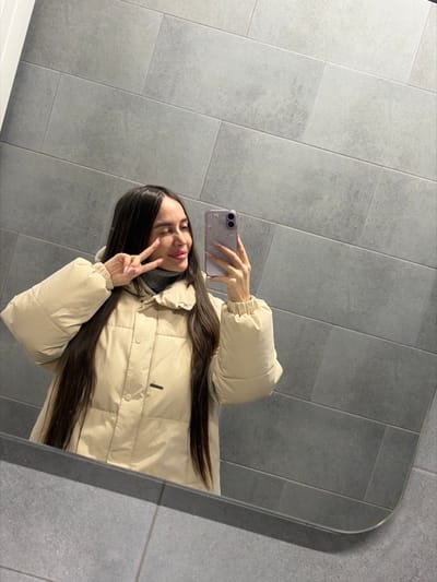 A single female individual is depicted in a mirror selfie within an indoor setting. The subject has long, dark hair and is wearing a light beige puffer jacket over a grey turtleneck. Her left hand is raised, forming a V-sign. Her right hand holds a light purple smartphone with a decorative case, positioned to capture her reflection. The individual exhibits a slight smile or smirk with partially closed eyes. The background consists of a wall covered in large, rectangular, dark grey tiles arranged horizontally. A tiled floor is partially visible in the reflection at the bottom of the mirror. The image contains no visual information to determine a specific location within the city of Novosaratovka, Russia.