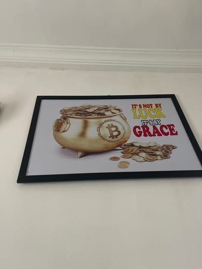A framed poster hangs on a white wall. The poster depicts a gold piggy bank with the Bitcoin symbol, overflowing with coins. Above the piggy bank, the poster reads, "It's Not by Luck, It's by Grace." The location appears to be indoors, and there is no evidence indicating the city of Naze, Nigeria.