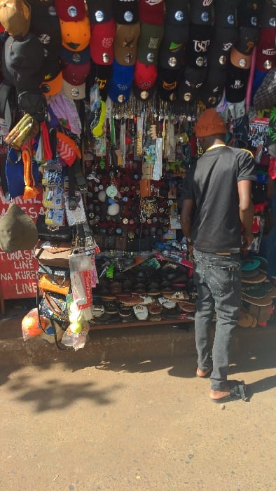 The image displays an outdoor market stall in Moshi, Tanzania, under bright daylight. The stall, filling the upper portion of the frame, is densely packed with merchandise. The top section showcases multiple rows of baseball caps and beanies, many displaying various logos and circular reflective stickers. Below these, a vertical display features numerous small items including keychains, watches, necklaces, and small bags, often against a red background. The bottom section of the stall consists of a multi-tiered rack holding numerous pairs of sandals and open-toed shoes. On the left side, a partial red sign with white text, reading "TUNASA LINE" and "NA KUREN LINE," is visible. An olive-green beanie hangs nearby.
An adult male, viewed from behind, stands facing the merchandise. He wears a dark short-sleeved t-shirt with a yellow stripe on the shoulder, dark grey jeans, and an orange knit cap. He is barefoot, standing on a reddish-brown, unpaved, and uneven ground surface composed of dirt and small stones. Shadows, indicative of strong sunlight, are cast onto the ground. A small, light-colored stone and a dark object lie on the ground near his feet. The man appears to be observing the items on display.