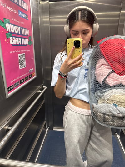 A woman is taking a selfie in an elevator with a yellow phone. She is wearing headphones and a light blue shirt with gray sweatpants. She is holding a laundry basket full of clothes. A promotional sign is visible on the elevator wall with an offer to "WIN YOUR FEES." The sign also contains a QR code. The elevator is located in a shopping centre in the city of Hull, United Kingdom.
