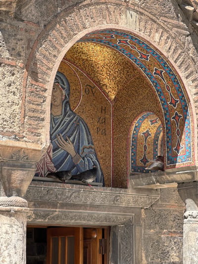 This media file depicts a close-up view of an ancient mosaic located on the exterior of a building, likely a church or a historical site, in Athens, Greece. The mosaic, framed by rough, weathered stone and brickwork, features a depiction of a reclining figure, presumably a religious icon, adorned in blue and white robes. The figure's face is visible, showing a serene expression.
The mosaic is characterized by its intricate tesserae, primarily gold, blue, and white, forming the figure and decorative borders. Surrounding the main figure, geometric patterns and stylized floral motifs are rendered in vibrant colors, including red and blue. Greek lettering is visible within the mosaic, though it is partially obscured and difficult to decipher in full.
Two dark-colored birds are perched on the stone elements to the right of the mosaic, appearing to interact with the artwork. The lighting suggests it is daytime, with shadows cast across the stone, indicating a bright, possibly sunny, day. The weathered texture of the surrounding stone and brickwork, along with the style of the mosaic, points to a location with significant historical and architectural importance in Athens. The overall impression is one of ancient artistry and enduring cultural heritage.