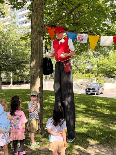 This media file captures an outdoor event likely taking place in Toronto, Canada, during the daytime under a bright, sunny sky with dappled shade from trees. The primary subjects are a performer dressed as a clown, standing on stilts and juggling three white balls, and a group of children watching him.
The performer is wearing a red vest over a white shirt, a red hat, and black and white striped pants. He has a red pouch attached to his belt. The children are dressed in casual summer clothing. One child, wearing a checkered shirt and green hat, is looking directly at the camera with an expressive face. Another child in the foreground wears a pink t-shirt with cartoon characters and the text "Morning Ritual" printed on it.
The scene is set in a park-like environment with a large tree in the foreground, providing shade. In the background, there is a street with a dark-colored car, suggesting the park is near a public road. A modern building with many windows is visible behind the trees. Colorful triangular flags and some signage, including a QR code and a sign with text that appears to say "MR. SPAGHETTI LEGS," are strung up near the performer, indicating some form of organized event or performance. The overall mood seems lighthearted and enjoyable, with the children appearing engaged and curious about the juggling act.