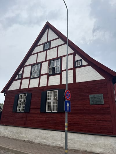 A historic two-story gable-ended building with a red and white facade stands prominently under an overcast sky in Liepaja, Latvia. The upper story features a distinctive half-timbered construction with white plaster infill framed by dark red timber beams. This section includes five windows: two larger rectangular windows on the main level, one of which has its upper casement open; one smaller square window on the far left; and two very small square windows near the apex of the gable, one directly at the top and another slightly below and to the right. The lower story is clad in horizontal red wooden planks, exhibiting a weathered texture. This lower section contains two large rectangular windows with white frames and closed black shutters. A dark, rectangular commemorative plaque is affixed to the red wooden wall on the right side of the lower story. The plaque bears text in both Latvian and Russian, indicating that Peter I (Peter the Great) lived in this house in 1697. The building rests on a textured white foundation, which meets a cobblestone street or sidewalk. A gray metal street pole is positioned in front of the building, slightly to the left of center, bearing two signs: a circular "No Stopping or Standing" sign (red diagonal line on a blue circle within a red border) above a blue rectangular "Straight Ahead" arrow sign pointing upwards. The sky is uniformly gray and cloudy, indicating an overcast day.