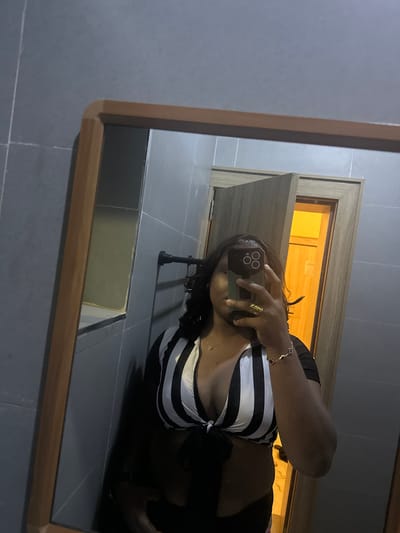A woman is taking a mirror selfie in what appears to be a bathroom. She is wearing a black and white striped crop top tied at the front and dark bottoms. She has dark hair and is wearing a gold ring and bracelet. The bathroom has grey tiled walls and a wooden framed mirror. A door is visible in the background, slightly ajar, with a warm light coming from the room beyond. The overall impression is of a casual, personal moment captured in a private space.