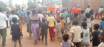 This image captures a crowded outdoor scene on an unpaved, reddish-brown dirt path within the city of Fizi, Democratic Republic of the Congo. Numerous individuals, predominantly children and young adults of African descent, are walking in various directions, largely away from the camera. Several people are engaged in carrying items; notably, multiple red and orange plastic basins are balanced on heads, with at least one red basin containing green leafy produce. A child in the foreground, left, holds an orange bucket. In the mid-ground, a person wears a yellow t-shirt and red pants, and another wears a red top and red pants while carrying an orange basin on their head. Further in the background, a person is visible wearing a red uniform with white text and a white helmet. On the right side of the frame, a man in a blue cap and green shirt is seated on a red plastic chair next to a brick building. The environment includes simple structures with brick walls and corrugated metal roofs lining the path, and distant green vegetation under diffused natural light.