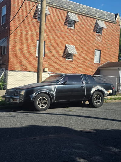 The image shows a dark-colored, two-door coupe parked on a street in front of a brick building. The car appears to be older and shows signs of wear, with rust and missing paint visible on the body. It has aftermarket wheels. The building has multiple windows with striped awnings. A wooden utility pole stands in front of the building. The street is paved and has a manhole cover. The scene is outdoors, under a clear sky. The location is identified as the city of Philadelphia, United States of America.