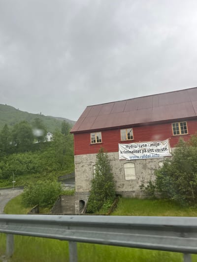 The image shows a red-roofed building with a concrete lower section, set against a cloudy, overcast sky. A white banner with blue text is attached to the side of the building. The banner reads "Hydro/ Lyse - miljø- kriminalitet på sitt verste. www.roldal.info". The building appears to be industrial or a large workshop, with several windows visible. Surrounding the building is lush green vegetation, with trees and a grassy hillside. In the distance, a small white house is visible nestled on the hillside. The scene suggests a rural or semi-industrial setting in Norway, possibly Røldal, given the text on the banner. The weather appears to be damp and cloudy, contributing to a somewhat somber mood. A grey metal guardrail is visible in the foreground, suggesting the image was taken from a road or elevated viewpoint.