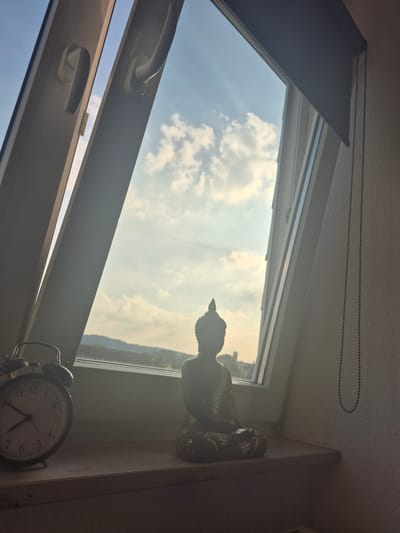 This is a still, medium shot taken at eye-level of a meditation scene, potentially in Hagen, Germany, viewed through a sloped window. The sky is visible through the window, with a few clouds and sunlight streaming in, suggesting late afternoon or early morning.

In the foreground, on a windowsill, sits a small, dark-colored Buddha statue in a meditative pose. To the left of the statue is an old-fashioned, silver-colored alarm clock with a white face and black numbers. The clock's hands point to approximately 10:10. The windowsill appears to be made of a light-colored wood or laminate.

The window frame is white, and the window itself is partially open, allowing a glimpse of the sky and a distant cityscape. The building outside the window has a textured facade. To the right of the Buddha statue, a dark string of beads hangs vertically against the wall, reaching from near the top of the frame towards the bottom. The overall lighting is soft and atmospheric, with some areas in shadow and others illuminated by the natural light. The scene evokes a sense of calm and introspection.