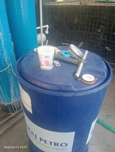 The image captures a scene featuring industrial or utility equipment, likely related to water treatment or storage, in what appears to be an outdoor or semi-enclosed area.

In the foreground, a large, cylindrical blue plastic drum (barrel) with a flat top dominates the lower right and center of the frame. A white label is affixed to its side, clearly displaying the text "RAJ PETRO" in large, dark letters. Below this, smaller text is partially visible, reading "MENTAG GROUP COMPANY".

The top of this blue drum serves as a temporary surface for various items. These include a white plastic cup with a floral design featuring pink and blue flowers and some illegible decorative text. Next to the cup are two small white rings, possibly washers or gaskets. Further to the right, a segment of a silver metal pipe, a roll of blue tape (likely plumber's thread seal tape), and a circular white component with a black handle (possibly part of a valve or pump) are resting on the drum's lid. A small orange-rimmed white cap, resembling a bung or closure, is also visible on the drum's top surface towards the right.

To the left of the main blue drum, two tall, upright blue cylindrical objects are partially visible. These appear to be larger tanks or filtration units, with a somewhat textured or streaked surface, possibly made of fiberglass or plastic. White PVC pipes and fittings are visible in the background, connecting to or running near these taller blue cylinders, suggesting a plumbing system.

Behind the equipment, a black mesh fence or netting is visible, forming the background of the scene. The ground beneath the objects appears to be concrete or a similar paved surface. A green hose is also visible on the ground towards the bottom right.

The image has a timestamp in the bottom left corner, indicating "2026/02/11 08:19", suggesting it was taken on February 11, 2026, at 8:19 AM. The lighting suggests it is daytime. No specific activity is depicted, but the arrangement of components on the drum implies a workspace or an area for maintenance and storage.