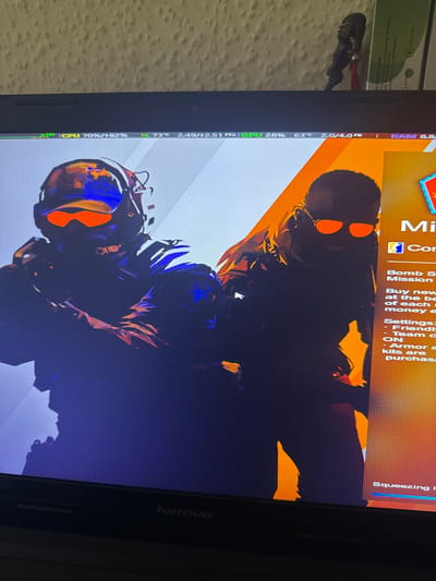 This media file shows a close-up of a laptop screen displaying a video game. The game appears to be a first-person shooter, with two character models prominently featured in silhouette. The characters are wearing futuristic combat gear, including helmets with illuminated visors in orange and blue. The background of the game screen is a vibrant orange and yellow, suggesting an abstract or stylized environment.

Visible on the left side of the laptop screen is a system monitor overlay, displaying real-time information about the laptop's performance. This includes CPU and GPU usage, temperatures (73°C and 63°C respectively), and RAM usage (2.0/4.0 GB). Text from the game's menu is also visible, with options like "Mission," "Bomb S," and "Settings" indicating gameplay elements.

The right side of the image shows the bezel of the laptop, with the "lenovo" logo clearly visible, identifying the device's brand. The overall scene is one of a person actively engaged in playing a video game on their laptop. The lighting suggests an indoor setting, likely a room at night or with dimmed lights, as the laptop screen is the primary light source. There are no obvious external environmental cues like weather or time of day beyond what's suggested by the indoor setting. The visible text, including system monitoring data and game menu options, provides specific details about the technical and gameplay aspects displayed.