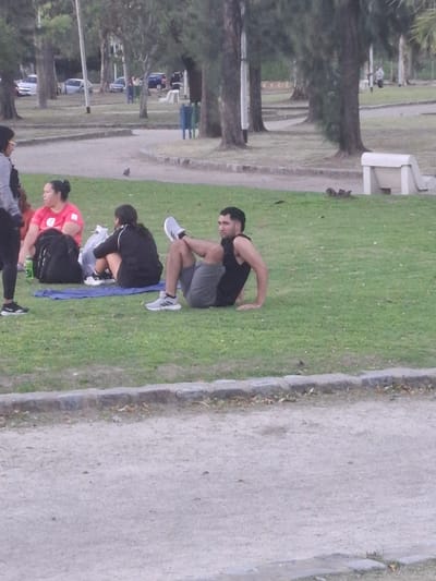 The image captures an outdoor scene in what appears to be a park or green space, with a group of people engaged in exercise or rest. The perspective is rotated 90 degrees, showing the scene as if viewed from above and to the side.
In the foreground, a man with dark hair and a beard, wearing a black tank top and grey shorts, is sitting on the grass in a squat-like position, with his hands resting on his knees. To his left and slightly below him, another person, dressed in a black long-sleeved top and black shorts/leggings, is seated on a blue mat, looking downwards, possibly stretching or preparing for activity. Beside this person, a woman in a red t-shirt with dark hair tied back is sitting on the grass, also looking down. Partially visible at the bottom left is another individual in dark clothing. A fifth person's face is barely visible at the top right, suggesting they are outside the main frame. All individuals appear to be wearing athletic shoes.
The setting includes a wide, light-colored paved or dirt path running alongside the grassy area. In the background, numerous trees line what appears to be another section of the park or a street, with hints of benches and possibly parked cars further in the distance. The lighting is soft and diffused, suggesting an overcast day or perhaps early morning/late afternoon. There is no visible text in the image. The general atmosphere is one of calm focus, characteristic of an outdoor exercise or relaxation session.