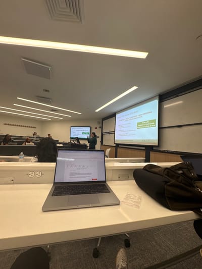 This image depicts a classroom or lecture hall scene at Babson College in Wellesley, Massachusetts, United States. The perspective is from a student's desk, looking towards the front of the room.

In the immediate foreground, a silver laptop is open on a white desk, displaying an interface titled "Application Question 1 of 5." To the right of the laptop, a small, clear plastic zip-lock baggie and a black backpack rest on the desk. Two electrical outlets are integrated into the desk's front panel. Below the desk, the feet of the person taking the photograph are visible, wearing light-colored sneakers.

In the midground, multiple students are seated in tiered rows of white desks. The heads and upper torsos of several individuals are visible, primarily with dark hair. A plastic water bottle is present on one student's desk.

At the front of the room, a female instructor, wearing a dark green top, stands behind a podium, actively gesturing with both hands towards the students. Behind her, two projection screens are visible. The main, larger screen on the right displays a presentation slide titled "Economic Order Quantity (EOQ) Model," detailing "Trade-offs: Variable vs. Fixed Costs" and "Risk Prone vs Risk Averse," with "BABSON COLLEGE" and "WWW.BABSON.EDU" at the bottom. A smaller screen to the left also displays presentation content, including a table or chart.

The classroom features a modern aesthetic with white walls, extensive linear fluorescent lighting integrated into the ceiling, and grey carpeted flooring. Ceiling vents are also visible.