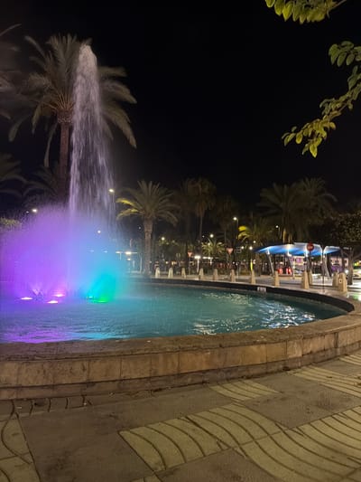 This media file captures a night scene at a fountain in Sant Antoni de Portmany, Spain. The fountain is illuminated with colorful lights, predominantly in shades of purple, blue, and green, creating a vibrant and magical atmosphere. Palm trees are visible, their fronds silhouetted against the dark sky, with one in the foreground being dramatically sprayed by the fountain's water jets.

The fountain is set within a paved area, with a stone-lined edge bordering the water. The paving stones have a distinct pattern. In the background, there are lights and structures suggesting a public space, possibly a promenade or a plaza, with hints of buildings and outdoor seating areas. The time of day is clearly night, as indicated by the dark sky and the artificial lighting. The weather appears calm, suitable for an outdoor evening scene. There is no visible text in the media file. The overall impression is one of a lively, illuminated evening display, characteristic of a tourist destination.