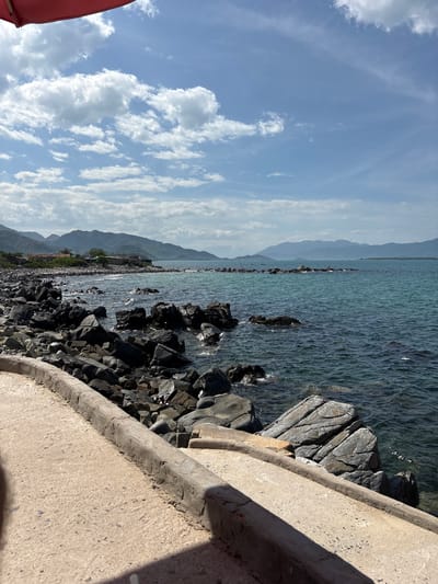 This is a vertical, eye-level shot of a rocky coastline and the ocean in Vĩnh Lương, Vietnam. The foreground features a concrete pathway with a raised, weathered edge that curves to the right. To the left of the pathway are numerous dark, irregular rocks, some of which are partially submerged in the water. The ocean is a clear turquoise color with moderate waves and some whitecaps. In the distance, a chain of mountains forms the horizon, with several small structures and trees visible along the shore. The sky is blue with scattered white, puffy clouds and a hint of a red awning in the upper left corner.