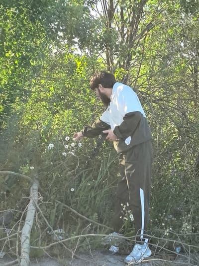 A man with a beard is standing in a wooded area, reaching out to touch some white flowers. He is wearing a white and dark green tracksuit and light blue and white sneakers. The setting appears to be a park or a natural area with lush green foliage and trees. Several fallen branches lie on the ground in the foreground. The lighting suggests it is daytime, likely late afternoon or early evening, with sunlight filtering through the leaves, creating a warm glow. The man's posture and focus on the flowers suggest a calm and contemplative moment. There is no visible text in the image. The location context is Ivanovo, Russia.