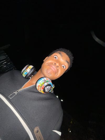 The image shows a young Black male wearing a black zip-up jacket with a white stripe and what appear to be JBL headphones decorated with stickers around his neck. The man is looking directly at the camera with wide eyes and a slightly distorted facial expression. The background is mostly dark, suggesting it is night, and the location may be Ilado, Nigeria, as requested. There is a faint light source above him.