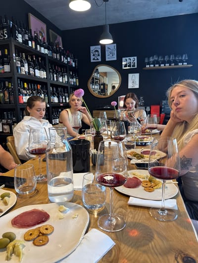 An indoor, slightly elevated, eye-level shot captures a group of individuals, primarily young women, seated around a light-colored wooden table at what appears to be a wine bar or restaurant, described as being in Vracar Urban Municipality, Serbia. The scene is dominated by the long wooden table, laden with multiple wine glasses, plates of food, and other serving ware. Several stemmed wine glasses, many containing red wine, are prominently displayed across the table. White ceramic plates hold various small food items including slices of cured meat, green olives, small crackers or pretzel-like snacks, and chunks of cheese. A clear glass carafe filled with water, along with smaller tumblers, are also present. White paper napkins are scattered on the table. A dark-colored cylindrical container, possibly holding cutlery or another item, is visible. A single purple dahlia flower is seen near one of the seated individuals. Five individuals are visible at the table. In the immediate foreground, to the right, a young woman with long blonde hair, wearing a light-colored top, rests her chin on her left hand, gazing towards the right of the frame with an introspective expression. A floral tattoo is visible on her left forearm. To her left, further down the table, a young woman with dark hair, clad in a white long-sleeved shirt, is looking downwards, seemingly at a mobile phone held in her hands. Behind her, another young woman with blonde hair, wearing a light-colored sleeveless top, is visible, looking slightly to her left. Her right hand is near her chin. Further in the background, to the right, another young woman with dark hair, wearing a dark top, is also observed looking at a mobile phone. A fifth individual, partially obscured, is visible on the far left side of the table, only their arm and shoulder are discernible. The background features dark, possibly black or very dark blue, walls. Extensive black shelving units, densely packed with numerous wine bottles of various shapes and labels, line the left and rear walls, indicating a specialized wine establishment. The ceiling appears to be concrete with suspended pendant lights. Several framed artworks and decorative items adorn the walls: on the left, an abstract piece with a pink background; on the right, multiple black-and-white minimalist prints depicting figures and objects (e.g., a person with a 'mystery' thought bubble, a drum set, multiple faces). An oval-shaped mirror with a wooden frame is mounted