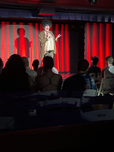 The image depicts a performance in Provincetown, Massachusetts, USA. On a stage, a person in drag, wearing a large black wig, heavy makeup, and a sequined top with a gold robe, holds a microphone and addresses an audience. The stage is set against a backdrop of red curtains, with stage lighting casting dramatic shadows, including a prominent silhouette of the performer on the left. The audience is seated in rows of chairs in the dimly lit foreground, with some individuals visible from behind. The overall atmosphere suggests an indoor entertainment venue.