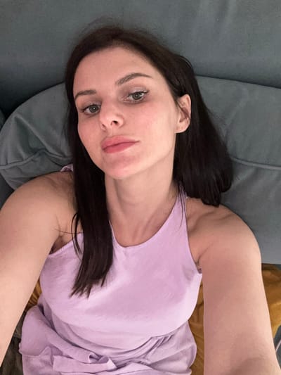 A woman with dark hair and light skin is lying on her side on a couch. She is wearing a light purple tank top. Her eyes are open and she is looking at the camera. Her lips are slightly parted. The couch appears to be upholstered in a dark gray fabric. A portion of a yellow pillow or blanket is visible behind her. The lighting suggests it is daytime. The overall impression is one of relaxation or informality.