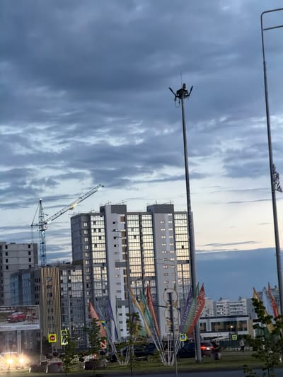 This is a vertically oriented photograph taken outdoors, likely in Nizhnekamsk, Russia. The scene captures an urban environment with multiple high-rise buildings, some appearing to be under construction or recently completed. A large construction crane is visible, with its arm extended towards the sky, suggesting ongoing building activity.
The sky is overcast with dramatic, textured clouds, hinting at a potentially unsettled or approaching storm, or perhaps the aftermath of one. Despite the cloudy conditions, there's a bright glow in the upper right portion of the sky, indicating either late afternoon or early morning light breaking through.
Several colorful flags or banners are strung along wires or poles, some bearing Cyrillic text. One banner clearly reads "САБАНТУЙ" (Sabantuy), which is a traditional Tatar festival. Another banner shows "САБАНТУЙ" as well. This suggests the photograph might have been taken around the time of this festival.
In the foreground, a tall pole extends upwards, with a device resembling a camera or sensor attached to it near the top. This could be part of a surveillance system, a weather monitoring station, or broadcasting equipment.
Roads and traffic are visible in the background, with cars present, including one with its brake lights illuminated. Street signs, including pedestrian crossing signs with yellow backgrounds and red circles, are also noticeable. The overall impression is a dynamic urban landscape undergoing development, with cultural elements (the banners) adding a specific context. The lighting suggests it's either dawn or dusk.