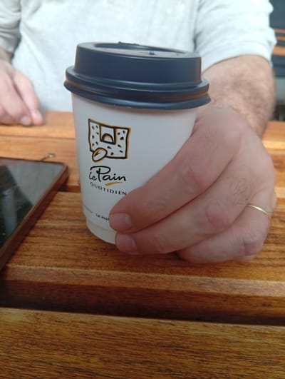 A medium shot depicts a person holding a paper coffee cup from "Le Pain Quotidien," a bakery chain. The cup is white with a dark blue lid and displays the bakery's logo. The person's hand, which has a gold wedding band on the ring finger, grips the cup. A portion of a smartphone rests to the left of the cup on a wooden table with a linear grain pattern. The person wearing a white t-shirt and light-colored long sleeve shirt. The location can be presumed to be near or inside the Aeropuerto Internacional Ezeiza (EZE) in Argentina, due to the "Le Pain Quotidien" branch there.