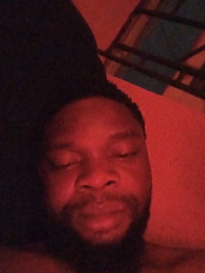 A close-up shot features an adult person, likely male, with dark skin and a short beard or heavy stubble, positioned centrally in the frame. The person's eyes are closed, suggesting they are sleeping, resting, or meditating. Their expression appears calm and relaxed.
The entire image is bathed in a strong, deep red light, giving everything a monochromatic, reddish hue. This red lighting highlights the contours of the person's face, emphasizing the nose, lips, and the texture of their beard.
On the left side of the frame, the background is a dark, indistinct mass, possibly a dark pillow, blanket, or fabric, providing a contrast to the illuminated face. On the right side and extending into the upper right corner, there is a reddish wall or surface, consistent with the overall red lighting. Faint, darker, geometric shapes, possibly part of furniture, a bed frame, or shelving, are visible in the upper right background, partially obscured by the dark red tone and grainy image quality. The overall setting appears to be an indoor space, likely a bedroom or a dimly lit room, with an intimate and subdued atmosphere created by the lighting. The image exhibits a slight graininess.