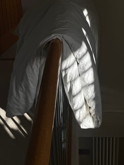 The image depicts a bedroom interior in a domestic setting, likely in Wealden, United Kingdom, though the exact location is impossible to determine definitively from visual cues alone. A white duvet or comforter is draped over a light-brown wooden bed rail. Sunlight filtering through what appears to be blinds or louvers casts distinct stripes of light and shadow across the bedding and the carpeted floor. The background shows a wall and what may be a heating radiator. No people are visible in the frame. The scene suggests a moment of undisturbed domesticity.