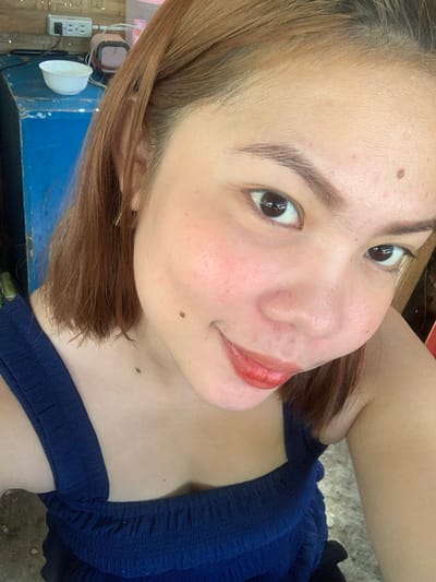 The image shows a close-up selfie of a young woman with light brown hair and fair skin, wearing a dark blue top. She is looking directly at the camera with a slight smile. Her makeup includes red lipstick and blush on her cheeks. There are a few moles visible on her face and chest. In the background, slightly out of focus, is a blue wooden surface, a white bowl, an electrical outlet, and some other objects that are not clearly identifiable. The image appears to have been taken indoors, possibly in a residential setting. There is no specific indication in the image to place it in Kiblawan, Philippines.