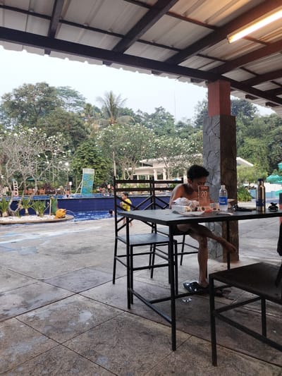 The image depicts an outdoor recreational area, likely part of a resort or water park, situated in Gunung Putri, Indonesia. The scene is viewed from under a covered patio featuring a white corrugated metal roof supported by dark brown metal beams and a structural pillar. This pillar has a reddish-brown upper section and a lower section clad in dark, irregular stone. A fluorescent light fixture is visible on the underside of the roof. The ground in the foreground consists of large, worn concrete tiles.
In the mid-ground, a blue-tiled swimming pool is the central feature. Various individuals, primarily children, are present in the pool, engaged in aquatic activities. On the left side of the pool, a decorative white rock and small plant bed includes a yellow fish-shaped water spitter. Further into the pool area, a blue climbing wall structure stands near the far edge. Beyond the pool, additional tables and chairs are visible, along with a white building and dense tropical foliage, including trees with white flowers (likely frangipani) and palm trees. The background is dominated by lush green trees under an overcast or hazy sky.
In the immediate foreground on the right, a young man with dark, curly hair is seated at a black square metal table. He is shirtless and wearing dark swim shorts, with his left leg crossed over his right. He appears to be examining something on the table. Objects on his table include a plastic bottle of "POCARI SWEAT," a dark insulated bottle, a stand-up sign displaying the number "20," and various white wrappers or napkins. A black and white sandal lies on the concrete floor near his feet. A young child in a bright yellow shirt is seen walking barefoot near the left edge of the swimming pool, facing away from the camera.