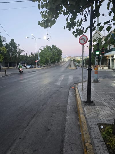 The image captures an intersection in Athens, Greece, likely during the early morning or late evening due to the soft twilight sky and illuminated streetlights. The scene is dominated by an asphalt road, wet from recent rain, with visible tire marks and some debris. A pedestrian crossing is marked on the road.
To the right of the road, a sidewalk paved with grey rectangular tiles is visible, bordered by a yellow curb. A black metal lamppost stands on the sidewalk. Adjacent to the lamppost is a traffic light, currently displaying a green signal for straight traffic and red for turning traffic. A circular red and white traffic sign with an arrow indicating "no right turn" is mounted on a pole nearby.
In the background, several trees with green foliage line the sides of the road. Buildings, some appearing to be shops or residences, are visible in the distance. A scooter rider wearing a helmet is seen moving away from the camera on the left side of the road, carrying a rectangular green sign on the back. Two pedestrians are standing near a parked car further down the road. In the far distance, hills or mountains can be seen under the pale sky, a common sight in Athens. The overall impression is a quiet urban street scene at a transitional time of day.