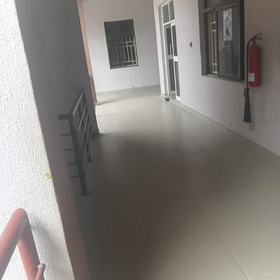 This is a photograph taken indoors, likely in a hallway or common area of a building. The image is oriented sideways, with the camera looking upwards. The scene features a red fire extinguisher mounted on a white ceiling. Below the ceiling, there's a large rectangular opening, possibly a window or a skylight, with visible internal framing. Further down, there's a smaller rectangular window with a metal grate on the outside. The walls appear to be a light color, possibly white or cream, and are tiled in large, rectangular sections. In the foreground, a textured white surface is visible, suggesting a wall or floor finish, and a portion of a red pipe or railing is present. The lighting suggests it might be daytime, but the indoor setting makes it difficult to determine the exact time or weather conditions. There are no people visible in the image. No legible text is visible.