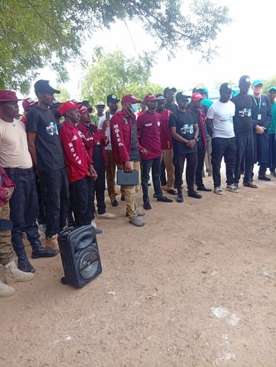 A group of approximately 20 people, predominantly young men, are standing in an outdoor setting, likely Dutse, Nigeria. They are gathered on a dirt ground, possibly a road or a clearing, with sparse vegetation and trees in the background. The sky is overcast, suggesting it could be a cloudy day.
The individuals are dressed in a mix of attire, with many wearing burgundy jackets or shirts with "NDLEA" printed on them, indicating affiliation with the National Drug Law Enforcement Agency. Some are also wearing baseball caps with similar branding or plain black caps. Several individuals are holding what appear to be tablets or small electronic devices. One person in the foreground is holding a black, portable speaker.
The scene suggests an official gathering or a public awareness event. The people appear attentive, possibly listening to someone or waiting for instructions. There is no clear indication of a specific time of day, but the lighting suggests daytime. The overall mood seems serious and organized.
Visible text includes "NDLEA" on the clothing and caps, and "DRUG ABUSE" on a t-shirt. The portable speaker has some text on it, but it is not legible. The ground is dry and dusty, with some footprints and scattered debris.