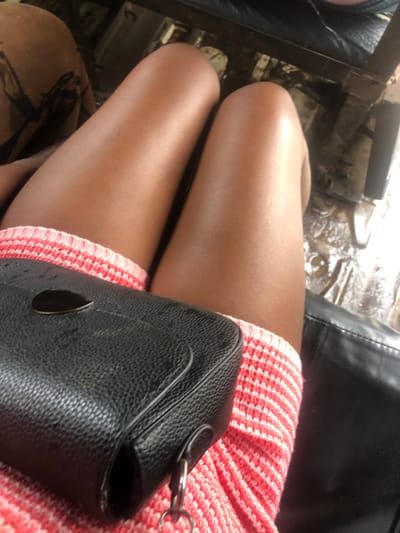 A high-angle, close-up photograph depicts the lower body of an individual seated within a vehicle in Ajah, Nigeria. Two bare, medium-dark skinned thighs are centrally positioned, with knees flexed. The individual is wearing a garment with a horizontal striped pattern in pink and white, visible at the upper thighs. A black, pebbled-texture handbag rests on the left thigh, featuring a silver-colored oval metallic closure on its flap and a silver D-ring attachment on its side. To the right of the individual's right leg, a dark, slightly reflective surface is visible. The vehicle's floor, visible beneath the knees, presents a dirty, irregular surface with standing water reflecting light, and dark structural elements. A third limb, clothed in brown patterned fabric, is partially visible in the upper left background.